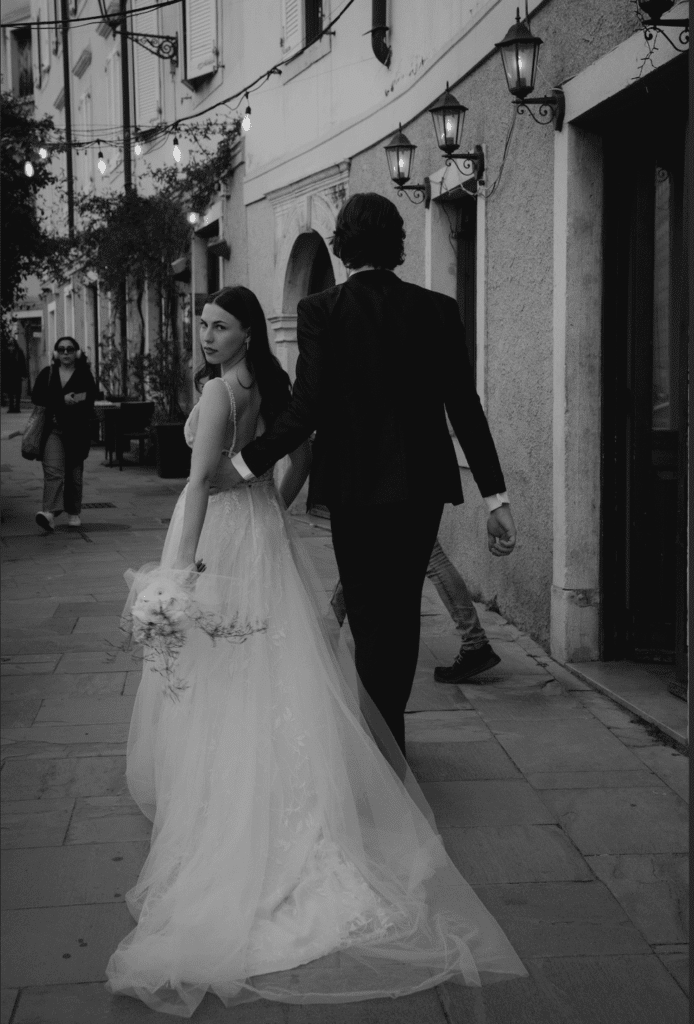 Ein Paar in formeller Kleidung geht Hand in Hand eine schmale Straße entlang und verkörpert damit die Essenz von Hochzeit. Die Frau, die ein langes weißes Kleid trägt, blickt über ihre Schulter. Dekorative Laternen hängen am Gebäude, im Hintergrund sind ein paar Fußgänger zu sehen. Schwarzweißbild.