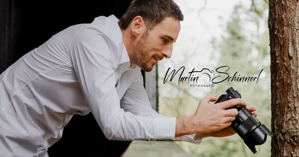 Ein Mann im weißen Hemd lehnt an einer Oberfläche und hält eine Kamera in der Hand, mit der er eine unvergessliche Hochzeit festhält. In der Nähe eines Fensters positioniert, ist er bereit, Momente zu verewigen. Im Vordergrund deutet das Logo „Martin Schinzel Fotografie“ auf sein Händchen für Hochzeitsplanung hin.