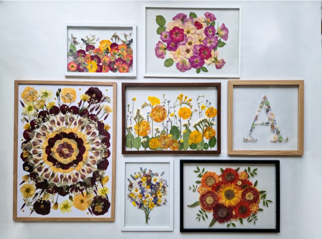 Eine Sammlung gerahmter Kunst aus gepressten Blumen schmückt eine weiße Wand, perfekt für Ihre Hochzeitsplanung. Die Rahmen variieren in Größe und Form und zeigen farbenfrohe Blumenmuster und einen aus Blumen gefertigten Buchstaben „A“. Kräftige Rot-, Gelb- und Lilatöne erwecken dieses einzigartige Hochzeitsdekor zum Leben.