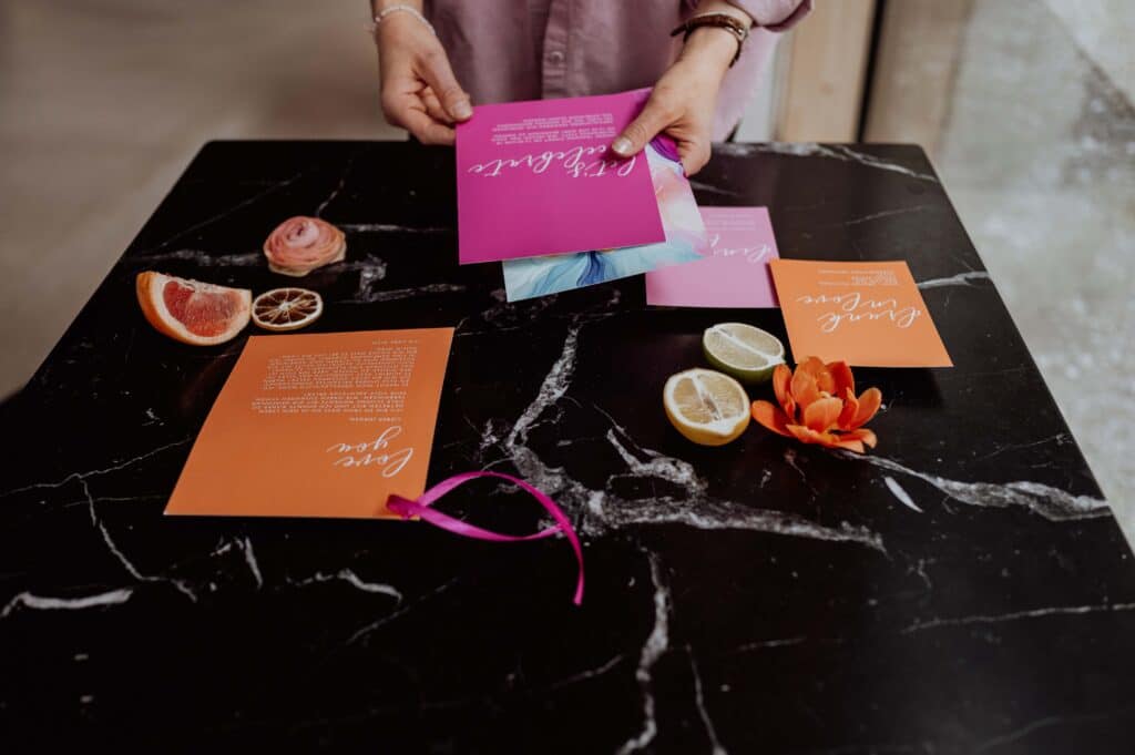 Eine Person arrangiert bunte Einladungskarten für eine Hochzeit auf einem schwarzen Marmortisch. Daneben liegen Grapefruit- und Zitronenscheiben, eine getrocknete Orange, eine rosa Ranunkel und eine orange Tulpe. Der Hintergrund ist sanft unscharf.