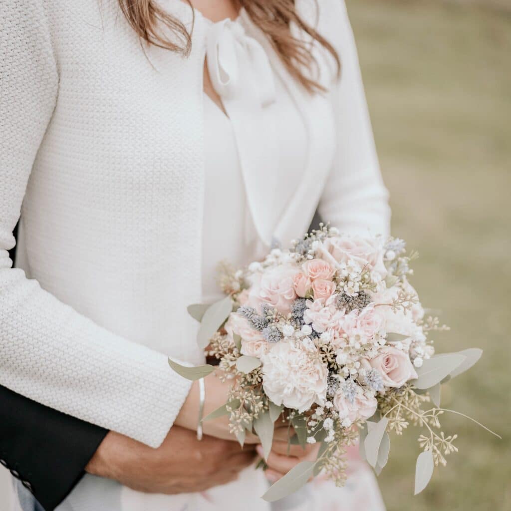 Eine Braut in einem weißen Outfit, umarmt von einer anderen, hält einen Strauß pastellfarbener Blumen inmitten üppigen Grüns. Die ruhige, grasbewachsene Kulisse unterstreicht diesen Moment des Hochzeitsglücks.