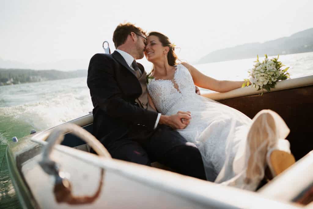 Ein Paar in Hochzeitskleidung sitzt an einem sonnigen Tag in einem Boot. Die Braut, die einen Blumenstrauß hält, lehnt sich zum Bräutigam hinüber, der einen dunklen Anzug trägt. Sie genießen ihren Hochzeitsmoment auf einem ruhigen Gewässer, eingerahmt von einer malerischen Kulisse, die perfekt für ein idyllisches Hochzeitserlebnis ist.