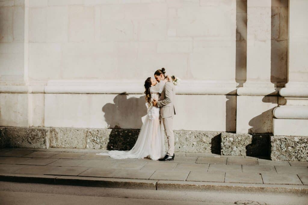 Ein Paar in Hochzeitskleidung küsst sich vor einem sonnigen Steingebäude im Hintergrund. Die Braut trägt ein fließendes weißes Kleid, während der Bräutigam einen hellen Anzug trägt. Ihre Schatten werden auf die Wand hinter ihnen geworfen – ein perfekter Moment, der ihr Hochzeitsglück einfängt.