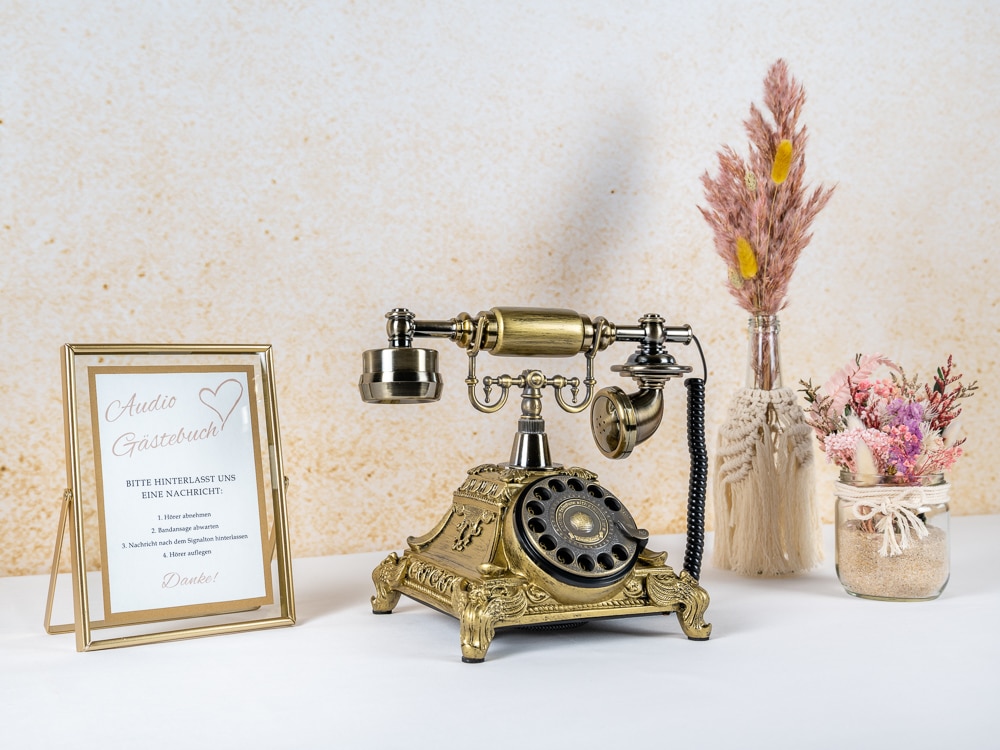 Ein Vintage-Wählscheibentelefon steht auf einem Tisch, umgeben von Hochzeitsdekorationen. Links ist ein gerahmtes Schild mit Text und einer Herzillustration, perfekt für jede Hochzeitsumgebung. Rechts schaffen getrocknete Blumen in einem Topf und einer Vase eine elegante, nostalgische Atmosphäre, ideal für die Hochzeitsplanung.