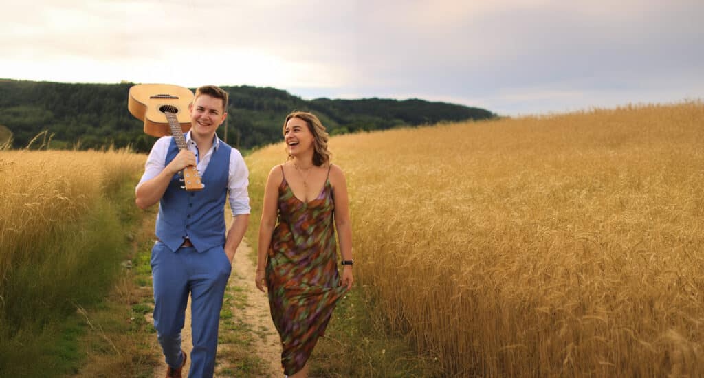 Ein Mann im blauen Anzug trägt eine Gitarre auf der Schulter und geht mit seiner lächelnden Braut im farbenfrohen Kleid spazieren. Sie schlendern gemütlich durch ein goldenes Weizenfeld, perfekt für Hochzeitsfotos, mit einem bewaldeten Hügel und einem bewölkten Himmel, die eine romantische Szene bilden.