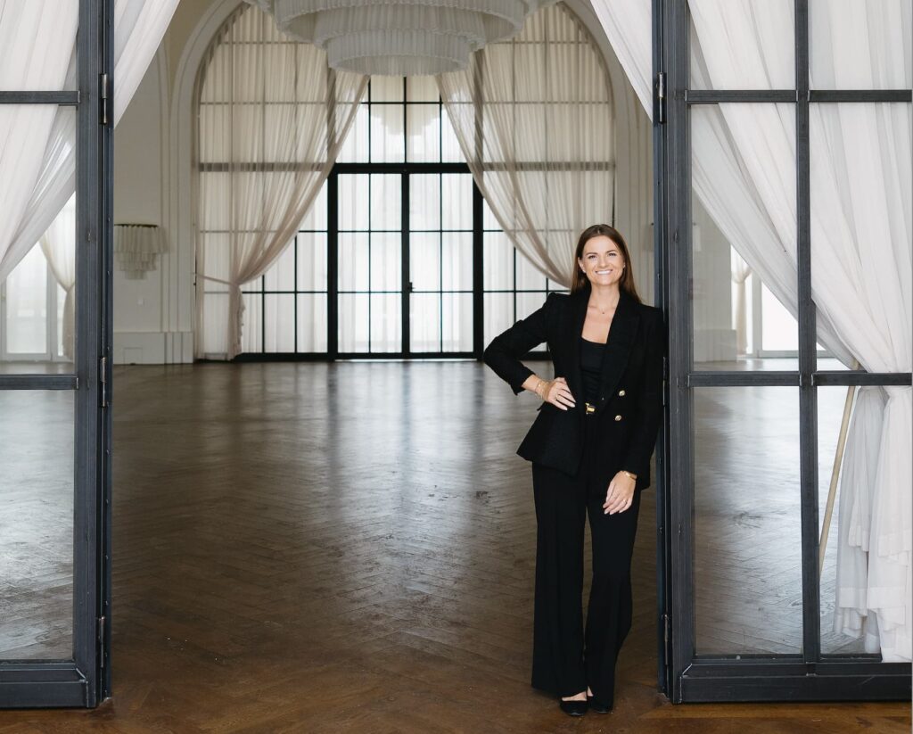 Eine Frau im schwarzen Anzug steht in einer offenen Tür und lächelt herzlich. Hinter ihr befindet sich ein eleganter, für die Hochzeitsplanung konzipierter Raum mit großen Fenstern, weißen Vorhängen und Holzböden.