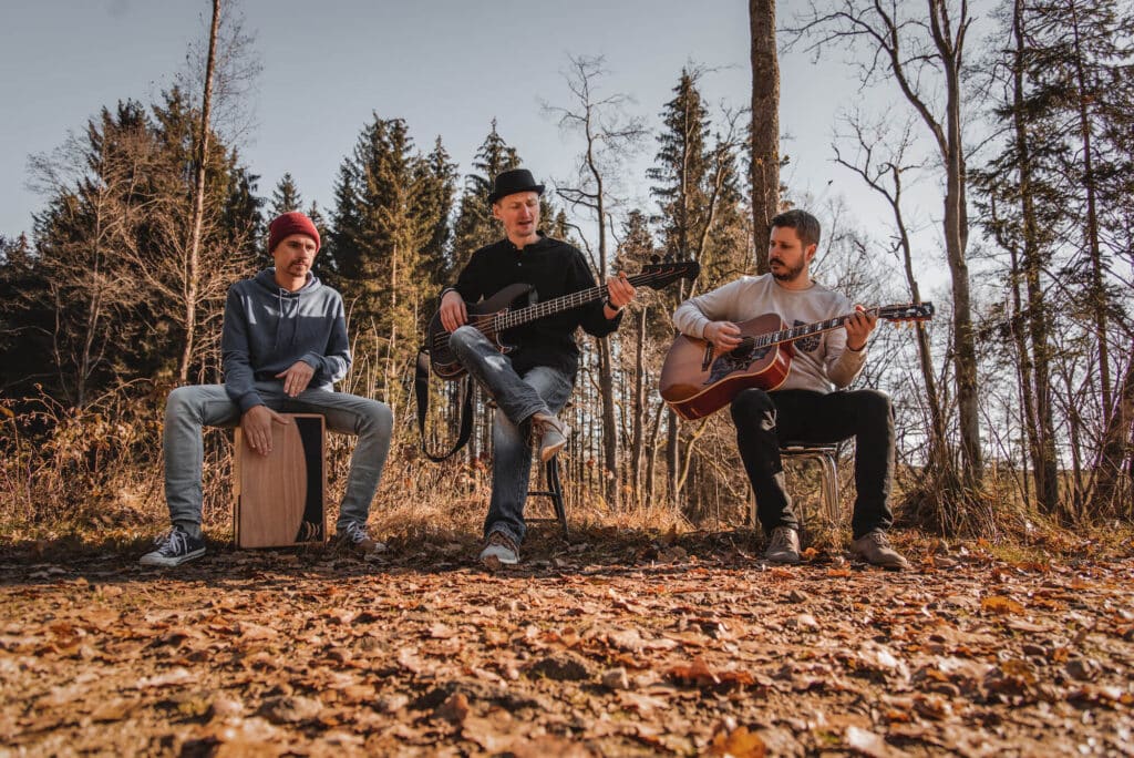 Drei Musiker, perfekt für jede Hochzeit, spielen Instrumente auf einer Waldlichtung. Einer sitzt auf einem Cajón, ein anderer klimpert auf einer Akustikgitarre und der dritte spielt einen E-Bass. Der Boden ist mit Laub bedeckt und hohe Bäume bieten eine atemberaubende Kulisse, ideal für Hochzeiten.
