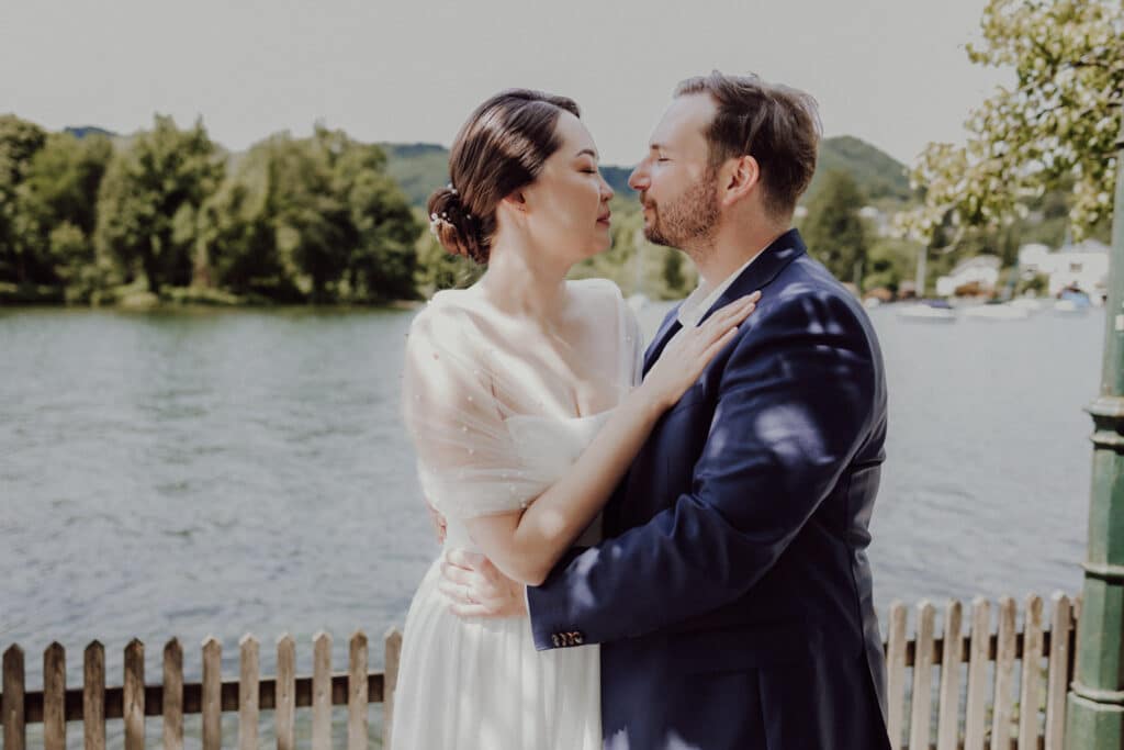 Ein Paar steht umarmend neben einem Holzzaun an einem See. Die Frau trägt ein weißes Kleid mit transparenten Ärmeln und der Mann einen marineblauen Anzug. Sie blicken sich vor einer malerischen Kulisse aus Wasser, Bäumen und Hügeln in die Augen.