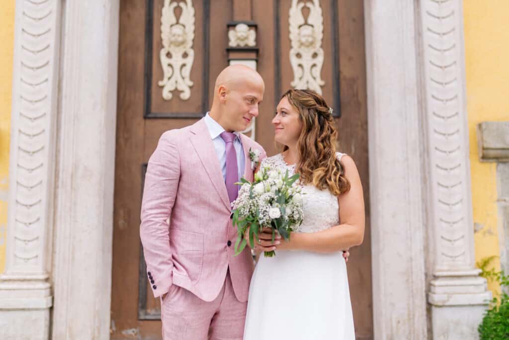 Ein Paar steht vor einer dekorativen Holztür. Der Mann trägt einen rosa Anzug und eine lila Krawatte, während die Frau ein weißes Kleid trägt und einen Blumenstrauß hält. Sie lächeln sich an und vermitteln einen freudigen und romantischen Moment.