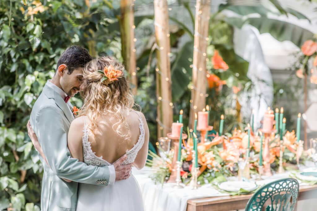 Ein Paar umarmt sich vor einem mit orangefarbenen Blumen und Kerzen dekorierten Tisch. Die üppige grüne Umgebung schafft eine romantische Atmosphäre. Die Frau trägt ein Haarteil mit Blumenmuster, der Mann trägt einen hellen Anzug.