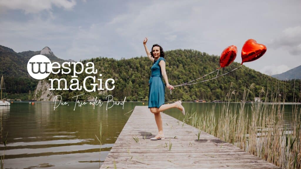Eine Frau in einem blauen Kleid steht auf einem Holzsteg an einem See und hält zwei rote herzförmige Luftballons in der Hand. Berge und Grün umgeben das Wasser. Der Text auf dem Bild lautet „Wespa Magic“ mit dem Slogan „Duo, Trio oder Band“.