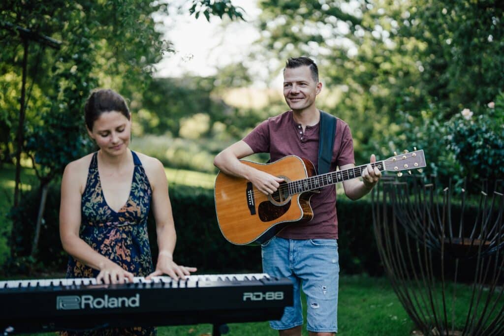 Eine Frau spielt ein elektrisches Keyboard, während ein Mann neben ihr steht und eine Akustikgitarre spielt. Sie befinden sich draußen in einem Garten, umgeben von Grün, und beide scheinen die Musik zu genießen.