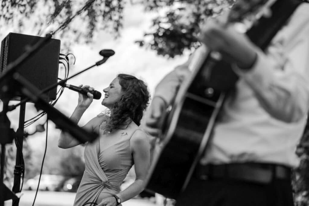 Eine Frau singt im Freien in ein Mikrofon, begleitet von einem Mann, der Gitarre spielt. Das Schwarzweißbild fängt ihren freudigen Gesichtsausdruck vor einem unscharfen Hintergrund aus Bäumen und Himmel ein.
