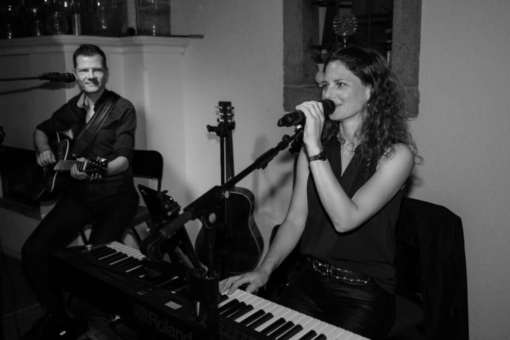 Schwarzweißbild einer Frau, die singt und Keyboard spielt, begleitet von einem Mann, der Gitarre spielt. Sie sitzen beide und sind in eine musikalische Darbietung vertieft, wobei ein Mikrofon und Instrumente sichtbar sind.