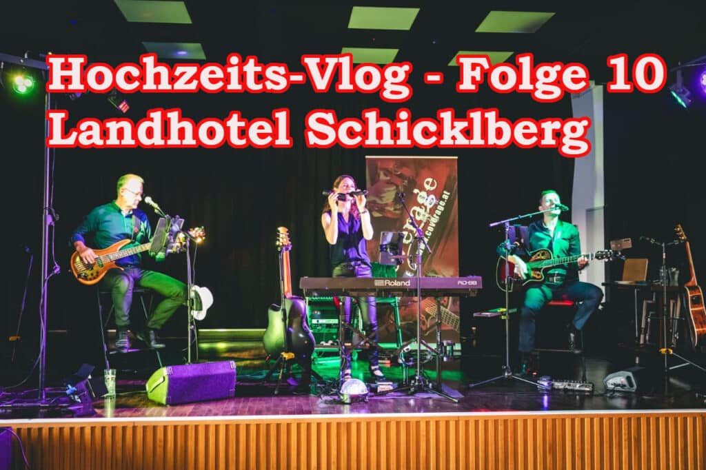 Auf der Bühne tritt eine Band mit Keyboard, zwei Gitarren und Mikrofon auf. Bunte Lichter illuminieren die Musiker. Im Hintergrund steht der Text: „Hochzeits-Vlog – Folge 10 Landhotel Schicklberg.“.