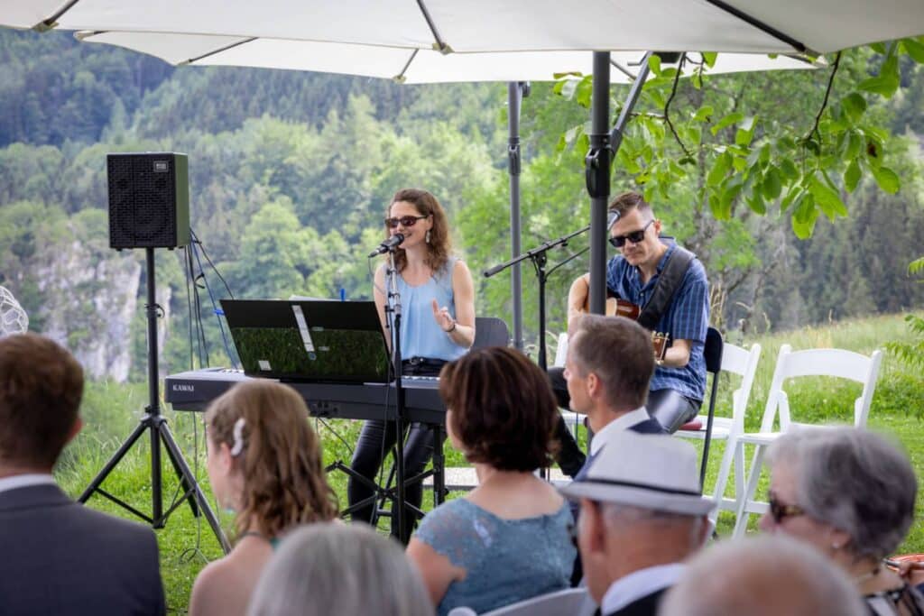 Eine Frau singt und spielt Keyboard, während neben ihr ein Mann unter einem weißen Sonnenschirm Gitarre spielt. Die Gäste sitzen auf weißen Stühlen und im Hintergrund ist eine üppige grüne Landschaft zu sehen.