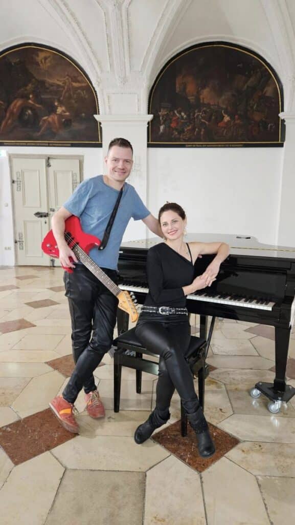 Ein Mann mit einer roten E-Bassgitarre steht neben einer Frau, die in einem eleganten Raum an einem Flügel sitzt. Beide lächeln. Der Raum hat gewölbte Decken und große Gemälde an den Wänden.