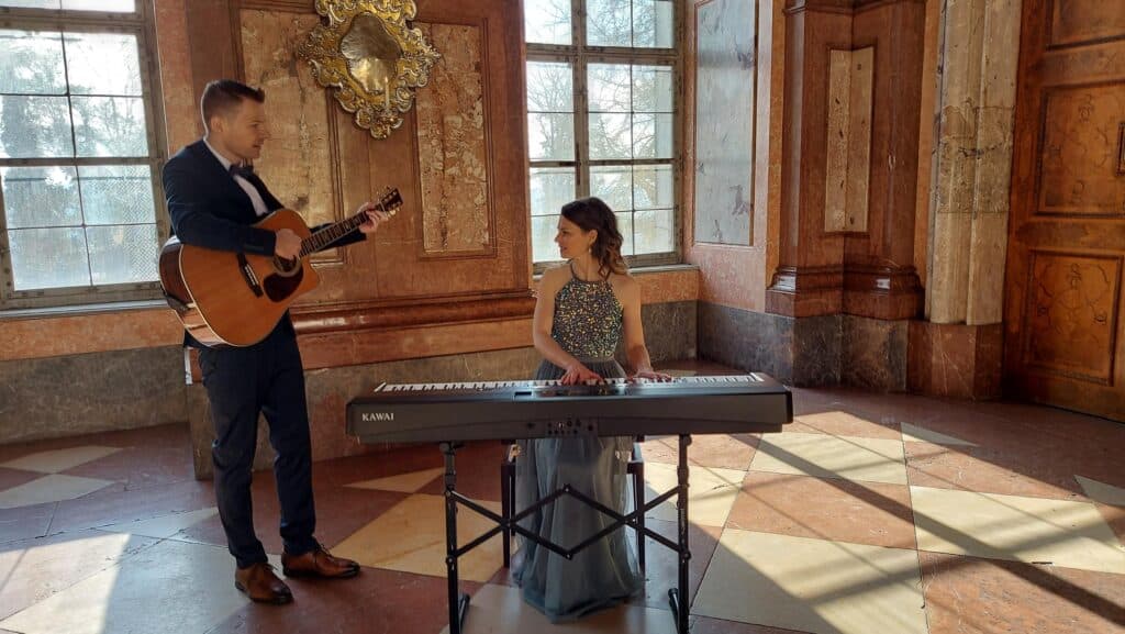 Ein Mann, der Akustikgitarre spielt, steht neben einer Frau, die an einem Keyboard sitzt. Beide spielen in einem sonnendurchfluteten, reich verzierten Raum mit großen Fenstern und Marmorwänden. Die Frau trägt ein glitzerndes Oberteil und einen langen Rock.