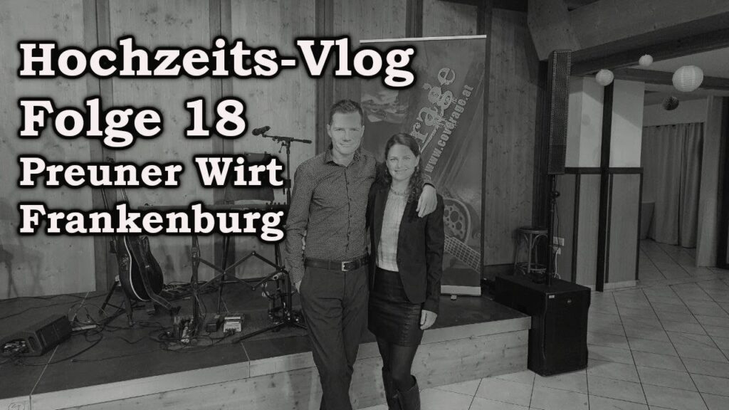 Ein Paar posiert vor einer Bühne mit Musikinstrumenten. Der Text lautet „Hochzeits-Vlog Folge 18 Preuner Wirt Frankenburg“. Der Veranstaltungsort hat Holzwände und einen gefliesten Boden.