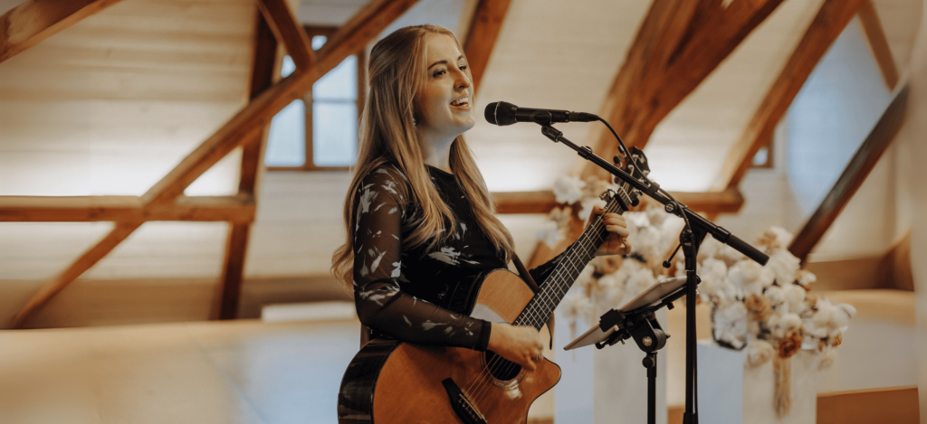 Eine Frau singt und spielt Akustikgitarre in einem warm beleuchteten Raum mit Holzbalken. Sie benutzt ein Mikrofon und steht vor einem Blumenarrangement.
