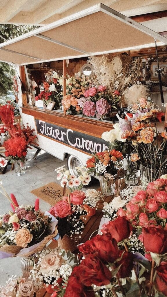 Ein farbenfroher Blumenwagen mit der Aufschrift „Blumenkarawane“, gefüllt mit einer Vielzahl farbenfroher Blüten, darunter Rosen, Nelken und Gänseblümchen. Die Auslage bietet satte Rottöne, sanfte Rosatöne und leuchtende Orangetöne und schafft so eine einladende und lebendige Marktszene.