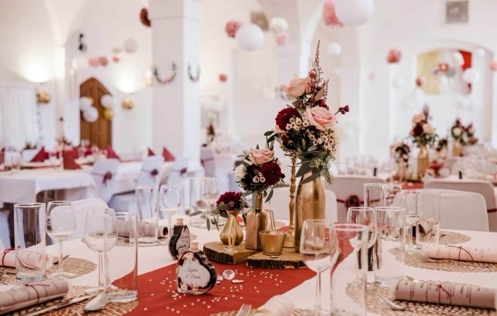 Ein wunderschön dekorierter Ort für Hochzeitsempfänge mit eleganten Tischen. Jeder Tisch ist mit goldenen Vasen mit rosa und roten Blumen ausgestattet, umgeben von Weingläsern, roten Tischläufern und verstreuten weißen Dekorationen. Sanftes Licht schafft eine warme Atmosphäre.