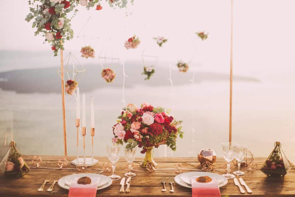Ein wunderschön gedeckter Esstisch mit elegantem Glasgeschirr, weißen Tellern und rosa Servietten. In der Mitte steht ein Tischdekor aus roten und rosa Blumen. Hängende Blumendekorationen und der Meerblick sorgen für eine romantische Atmosphäre.