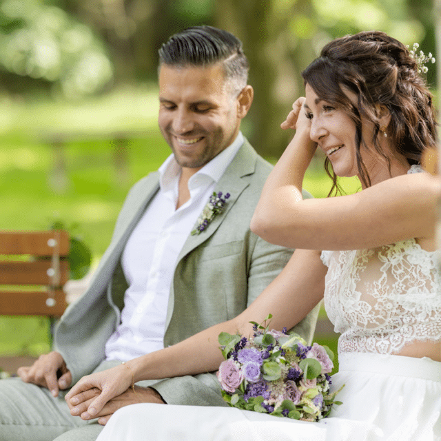 Ein lächelndes Paar sitzt draußen und hält sich an den Händen. Sie trägt ein weißes Kleid mit Spitzendetails und hält einen Strauß lila und weißer Blumen in der Hand. Er trägt einen hellgrauen Anzug mit einem weißen Hemd und einer kleinen Blumenansteckblume. Der Hintergrund ist grün und verschwommen.