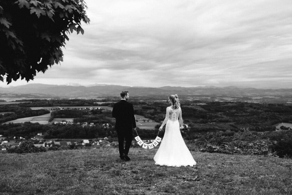 Braut und Bräutigam stehen auf einem grasbewachsenen Hügel und halten ein „Just Married“-Schild in der Hand. Ihnen blickt eine weite Landschaft mit sanften Hügeln und bewölktem Himmel entgegen. Die Szene ist in Schwarzweiß gehalten und schafft eine zeitlose, romantische Atmosphäre.