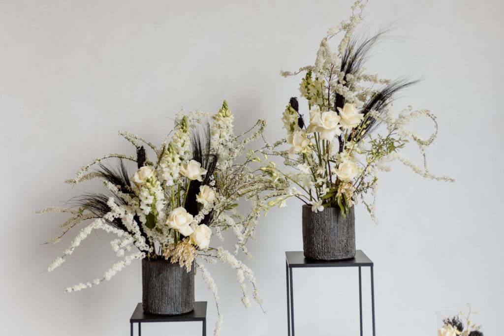 Zwei graue Vasen auf Ständern präsentieren elegante Blumenarrangements mit weißen Rosen, zarten weißen Federblumen und dunklen getrockneten Akzenten. Der Hintergrund ist eine schlichte, cremefarbene Wand.