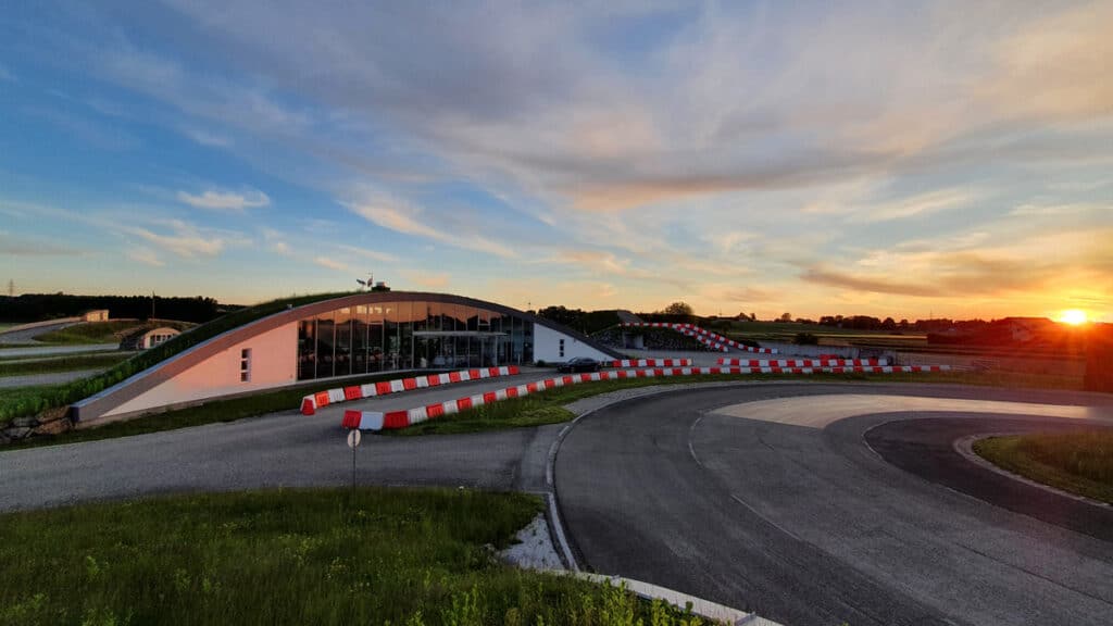 Eine kurvenreiche Rennstrecke, gesäumt von rot-weißen Barrieren, führt zu einem modernen Gebäude mit grünem Dach. Der Himmel ist mit dramatischen Wolken bedeckt, und ein Sonnenuntergang taucht die Landschaft in einen orangefarbenen Schimmer.
