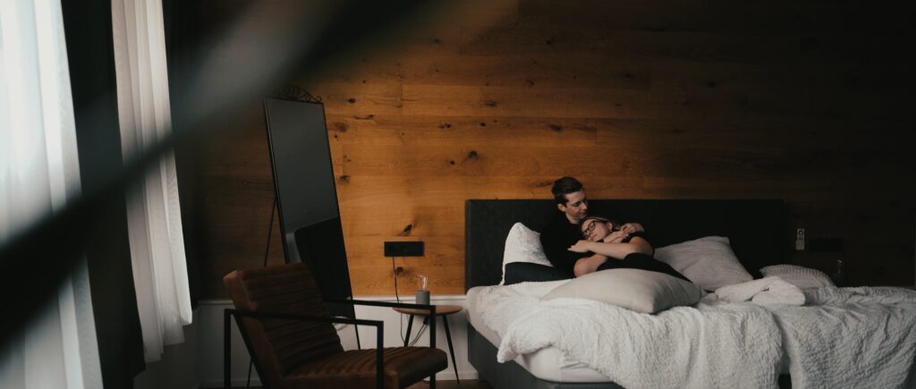 Ein Paar sitzt auf einem Bett in einem gemütlichen, schwach beleuchteten Zimmer mit Holzwänden. Sie kuscheln unter weißer Bettwäsche. Ein moderner Stuhl, ein Standspiegel und sanftes Licht schaffen eine warme Atmosphäre.