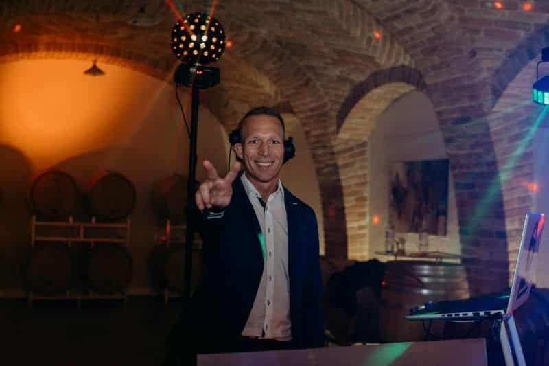 Ein lächelnder Mann mit Kopfhörern und Anzug steht hinter dem DJ-Equipment und zeigt ein Peace-Zeichen. Die Kulisse ist ein gemauerter Keller mit Weinfässern und warmer, stimmungsvoller Beleuchtung.