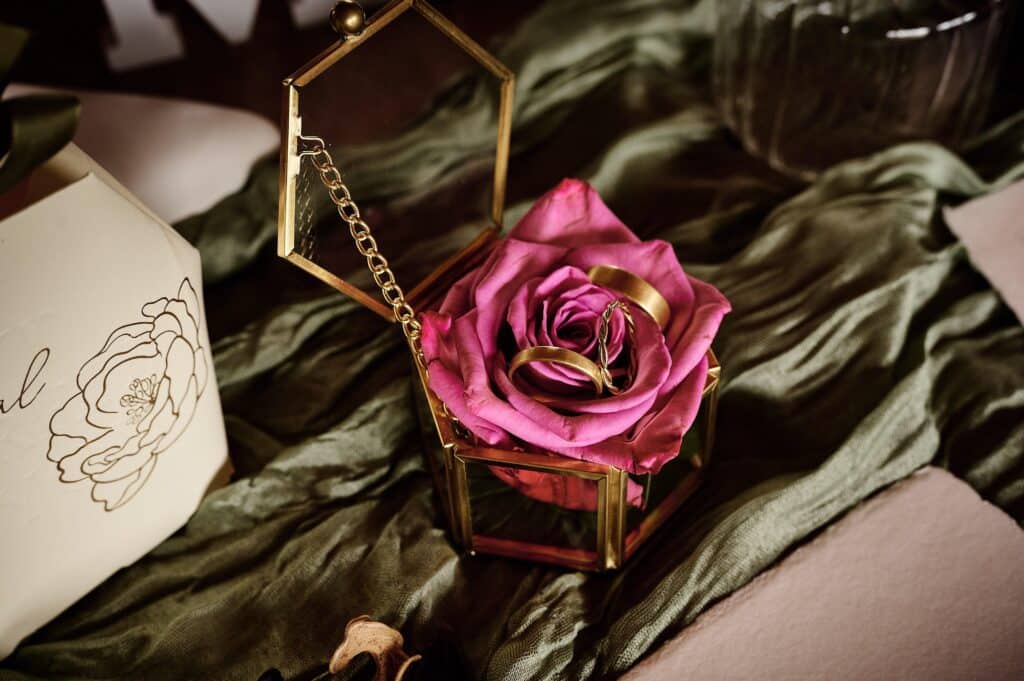 Eine goldene sechseckige Glasbox mit einer rosa Rose und zwei goldenen Ringen darauf, platziert auf einem grünen Stoff mit einer Blumenkarte daneben.