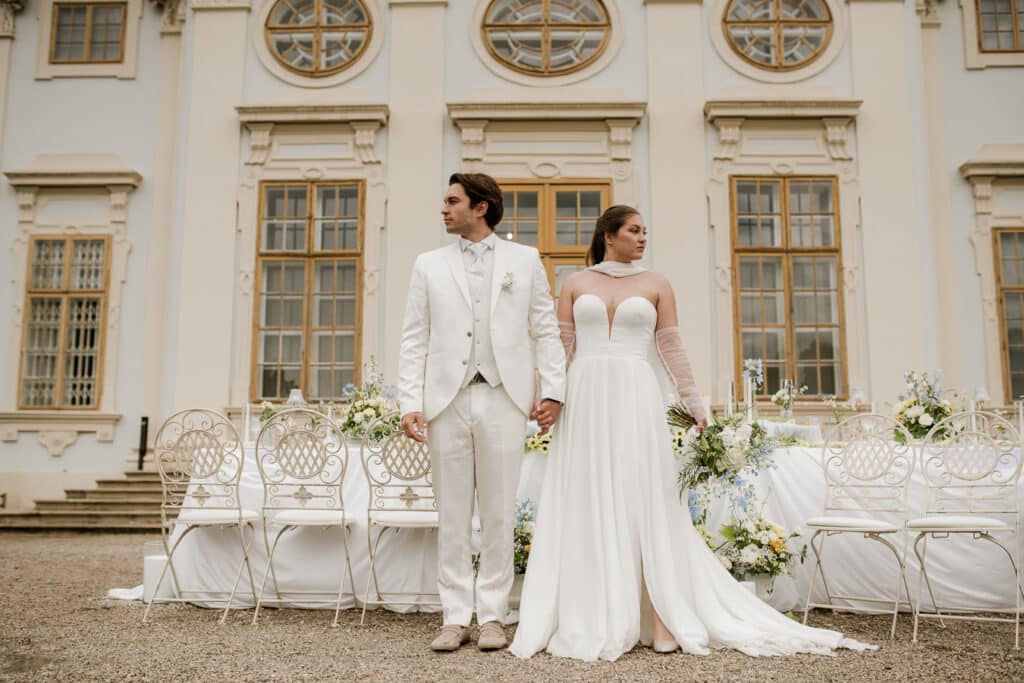 Eine Braut und ein Bräutigam in weißer Hochzeitskleidung halten sich an den Händen und stehen vor einem eleganten, verschnörkelten Gebäude und schauen in entgegengesetzte Richtungen. Hinter ihnen sind weiße Stühle und Blumenarrangements aufgestellt.