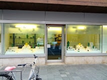 Eine Schaufensterfront mit großen Schaufenstern zeigt weiße Regale und Ständer mit verschiedenen Artikeln. Ein Fahrrad mit rosa Sitz ist auf dem Bürgersteig vor dem Ladeneingang geparkt.