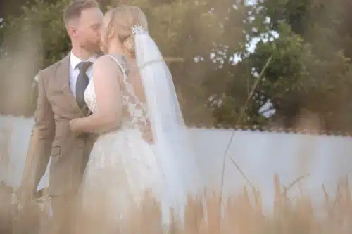 Eine Braut und ein Bräutigam küssen sich im Freien, umgeben von hohem Gras, Bäumen und einer weißen Wand im Hintergrund. Die Braut trägt ein weißes Spitzenkleid und einen Schleier; der Bräutigam trägt einen braunen Anzug und eine Krawatte.