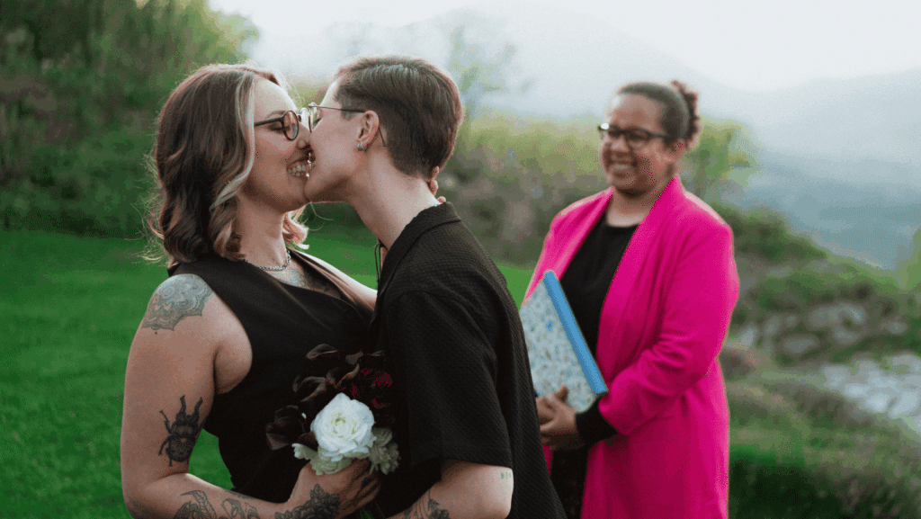 Zwei Personen küssen sich bei einer Hochzeitsfeier im Freien, halten Blumen und lächeln. Eine dritte Person in einer hellrosa Jacke steht hinter ihnen, hält ein Buch und lächelt. Im Hintergrund sind grünes Gras und Berge zu sehen.