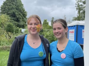 Zwei lächelnde Frauen in blauen Hemden stehen draußen. Die eine hat einen Vogel auf die Wange gemalt und trägt ein Abzeichen mit der Aufschrift "Luca". Die andere trägt ein Schild mit der Aufschrift "Lily". Im Hintergrund sind Grünpflanzen und eine mobile Toilette zu sehen.
