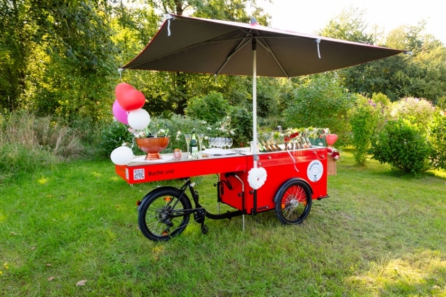 Ein roter fahrbarer Wagen mit einem Sonnenschirm steht auf einer Wiese und ist mit Luftballons und Blumen geschmückt. Der Wagen fasst Getränke und Gläser und ist umgeben von Bäumen und Grünpflanzen in einer parkähnlichen Umgebung.