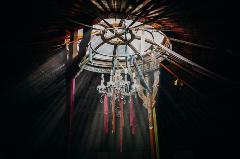 Das Sonnenlicht strömt durch das runde Oberlicht einer traditionellen Jurte und beleuchtet bunte, hängende Bänder und einen Kristalllüster im dunklen, hölzernen Innenraum.