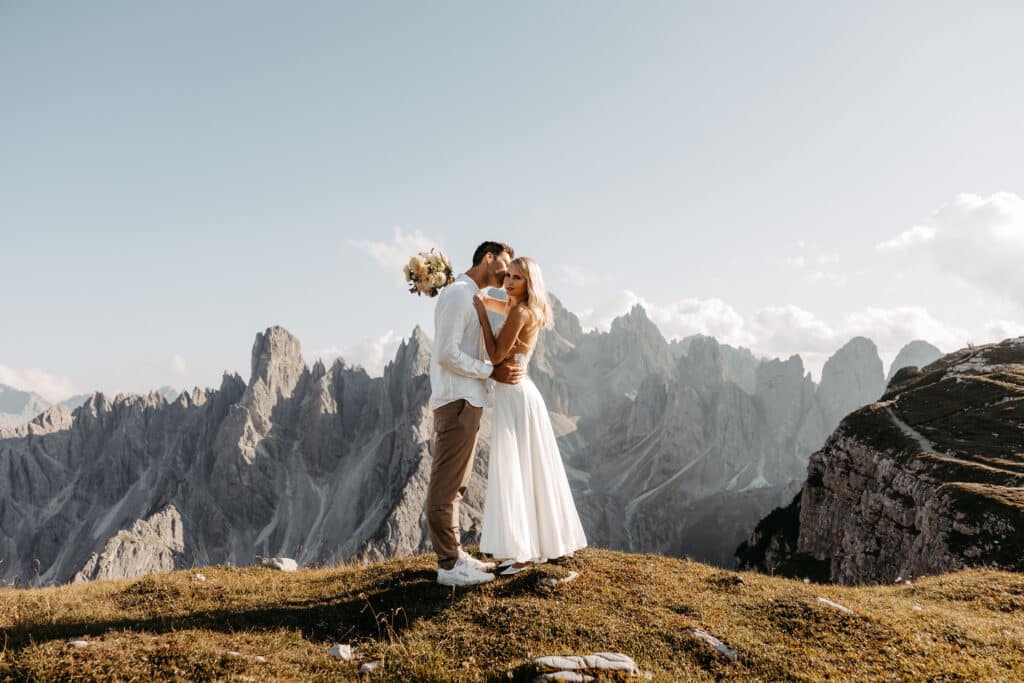 Ein Paar in Hochzeitskleidung steht auf einem grasbewachsenen Hügel, umarmt und küsst sich vor dramatischen Berggipfeln und einem klaren Himmel im Hintergrund. Der Bräutigam hält einen Blumenstrauß hinter der Braut.
