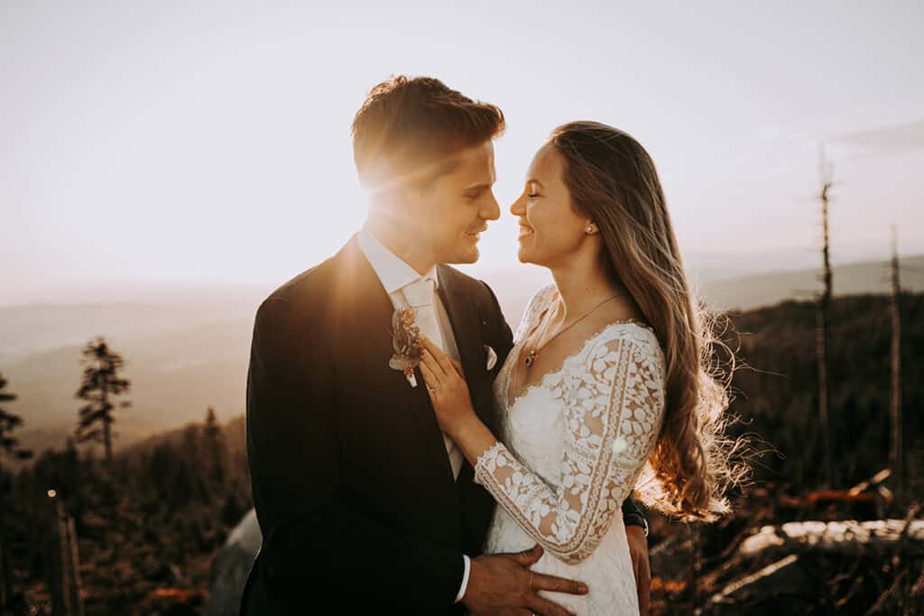 Eine Braut und ein Bräutigam stehen bei Sonnenuntergang eng beieinander, lächeln und blicken sich in die Augen. Die Braut trägt ein Hochzeitskleid aus Spitze und der Bräutigam einen Anzug. Im Hintergrund ist eine malerische, sonnenbeschienene Landschaft zu sehen.