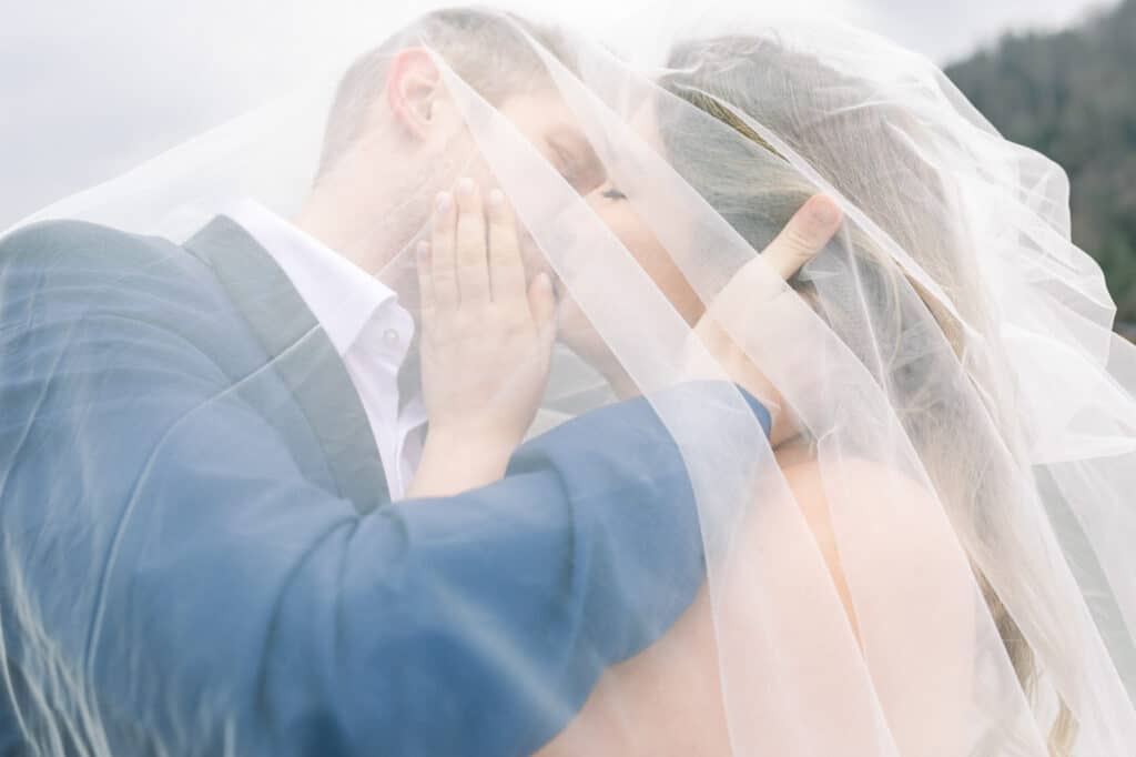 Eine Braut und ein Bräutigam umarmen sich und küssen sich unter einem weißen Schleier im Freien, wobei der Bräutigam sanft das Gesicht der Braut hält. Die Szene ist weich und romantisch, mit einem unscharfen natürlichen Hintergrund.