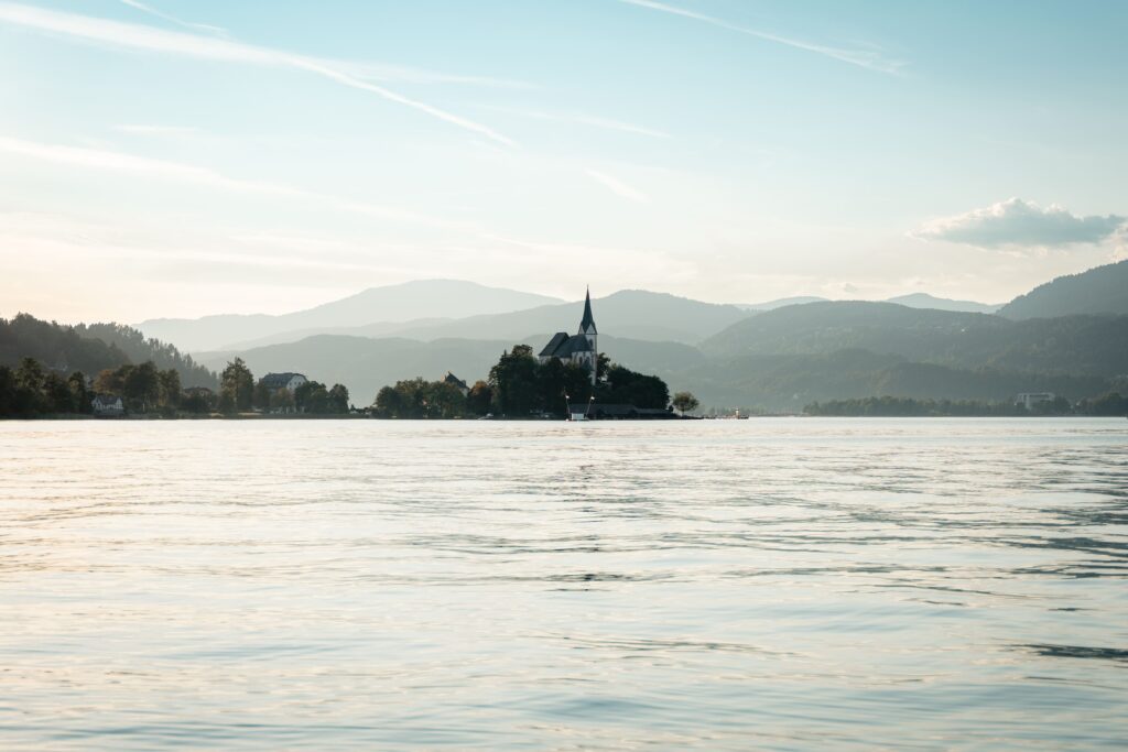 Ein ruhiger See mit sanften Wellen spiegelt eine Insel mit einer Kirche und Bäumen wider, umgeben von fernen Hügeln und einem sanften, blassblauen Himmel bei Tageslicht.