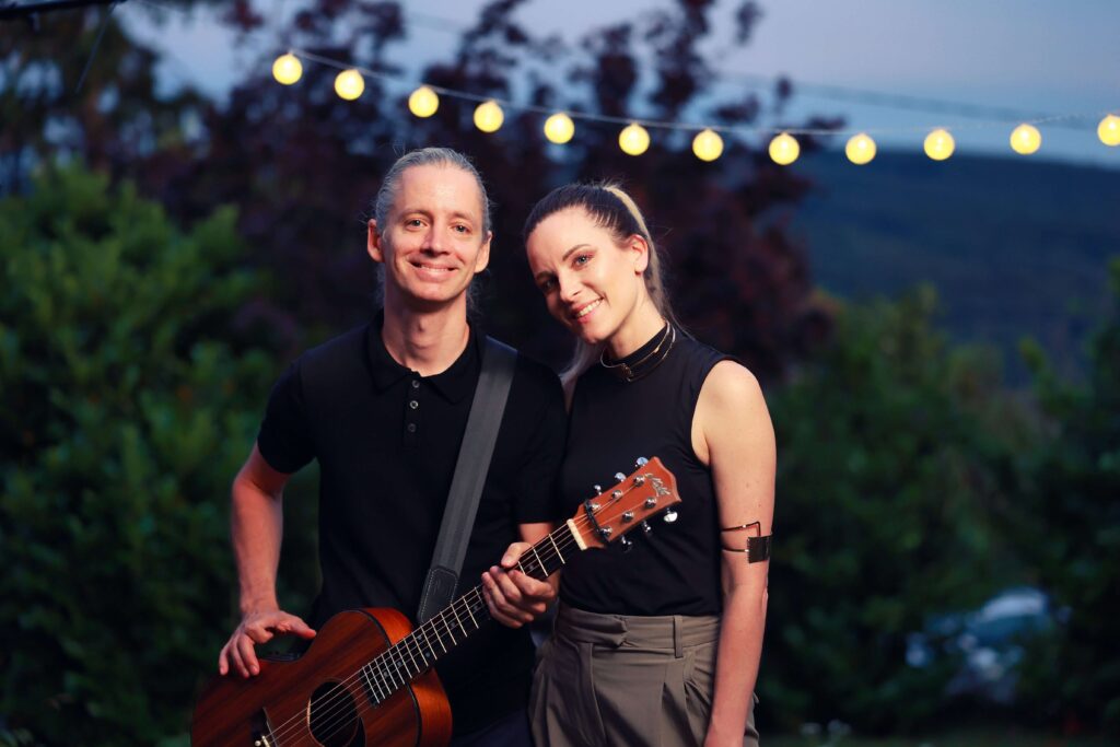 Zwei Personen stehen in der Abenddämmerung im Freien und lächeln in die Kamera. Einer hält eine Akustikgitarre in der Hand. Sie sind von Grün umgeben, über ihnen leuchten Lichterketten, und im Hintergrund sind Bäume und Hügel zu sehen.