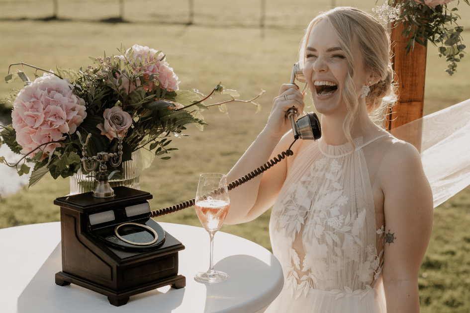 Eine lächelnde Braut im Spitzenhochzeitskleid lacht, während sie an einem mit rosa Blumen geschmückten Tisch im Freien ein altes Telefon in der Hand hält und ein Glas Roséwein in der Nähe hat.