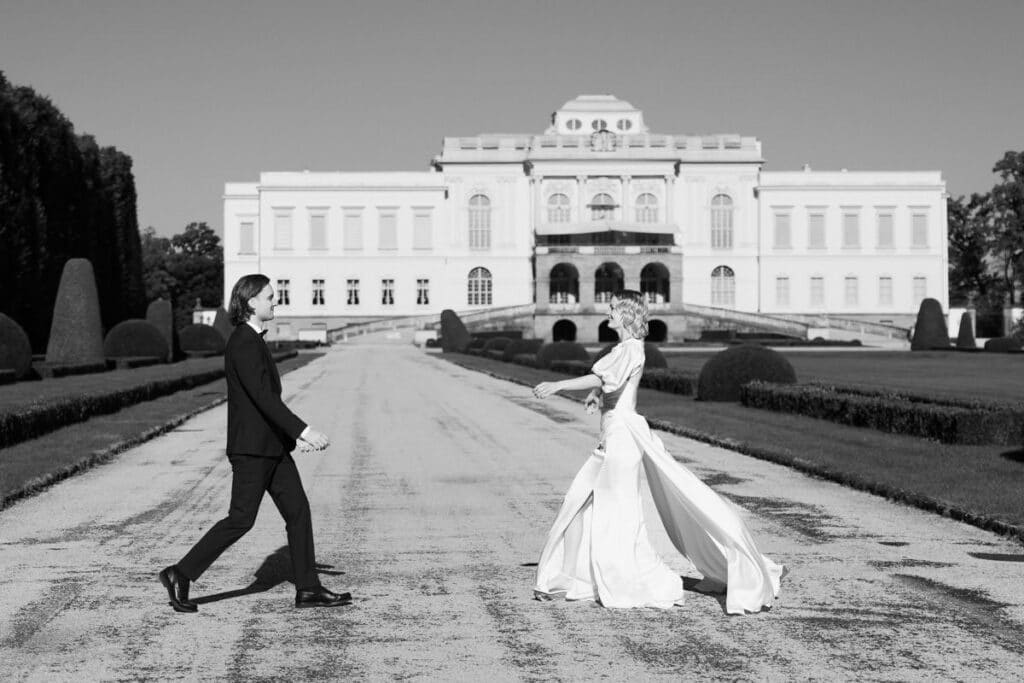 Eine Braut und ein Bräutigam gehen auf einem breiten Weg vor einem großen, eleganten Gebäude, umgeben von gepflegten Gärten, aufeinander zu. Die Szene ist in schwarz-weiß gehalten.
