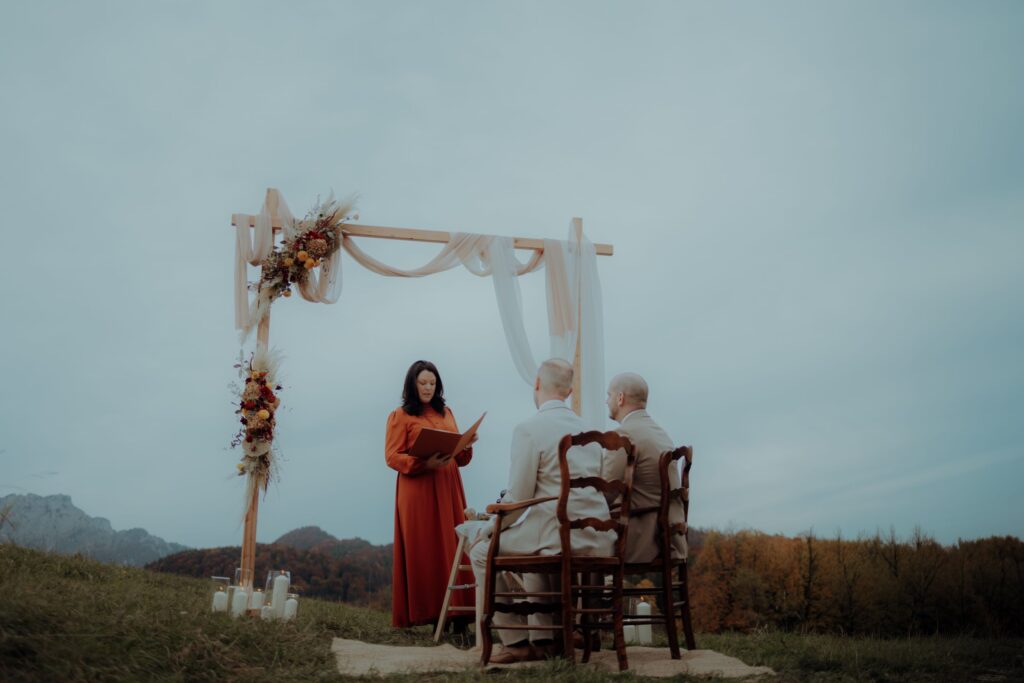 Eine Frau in einem roten Kleid liest unter einem verzierten Holzbogen im Freien aus einem Buch, während zwei Personen nebeneinander auf Stühlen sitzen und ihr gegenüber sitzen. Die Szene befindet sich auf einem grasbewachsenen Hügel mit Bäumen und Bergen im Hintergrund.