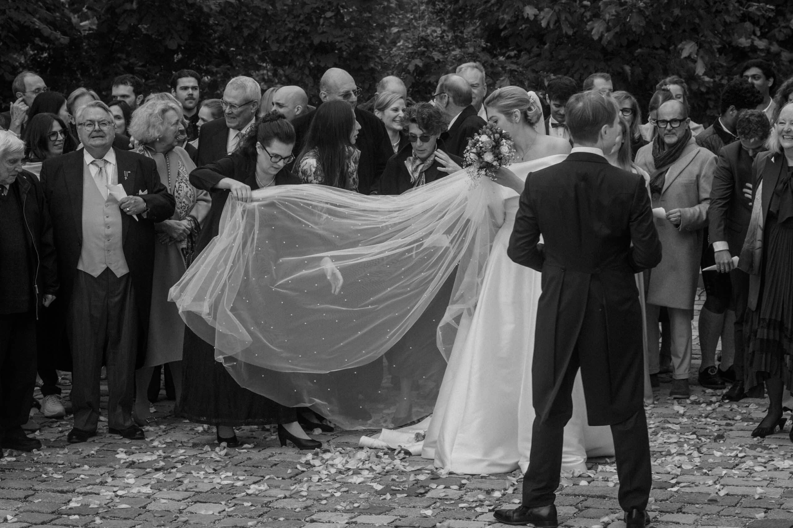 Eine Braut in einem weißen Kleid lässt sich ihren Schleier von zwei Frauen zurechtrücken, während eine Gruppe gut gekleideter Gäste zusieht. Die Szene findet im Freien statt, und die Stimmung ist feierlich. Blütenblätter werden auf den Boden gestreut.